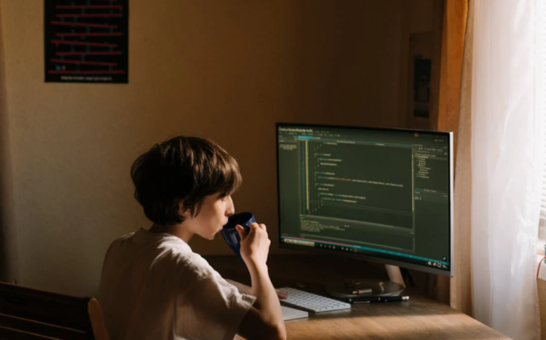 A person is seen sitting in front of a computer screen displaying complex programming code, indicating their engagement with a technical or computer-related task.